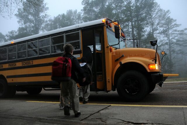 Children with backpacks board a yellow school bus on a foggy morning, parked by the sidewalk. Tall trees loom in the background, and "Parish Schools" is written on its side. Amidst this serene scene, it’s crucial to consider school bus accidents and who is liable for the safety of these young passengers.