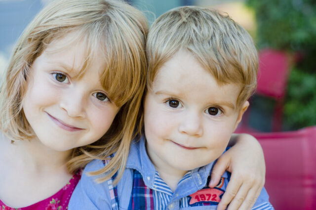 A young girl with blonde hair smiles and wraps her arm around a younger blonde boy with big brown eyes. They look directly at the camera and sit closely together, appearing happy and affectionate. The background is blurred with greenery and red elements.