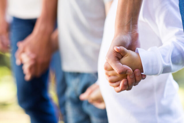 A close-up image of a group of people holding hands, forming a line. The focus is on the hands, showing a mix of adult and child hands lightly gripping each other. The background is blurred, with green and yellow tones suggesting an outdoor setting.