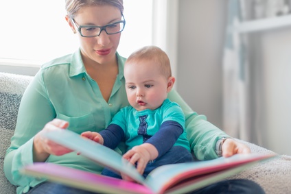 An adult wearing glasses reads a book to a baby who is sitting on their lap. Both are dressed in teal tops. The baby appears curious and is touching the pages of the book. They are sitting on a light-colored sofa in a bright room.