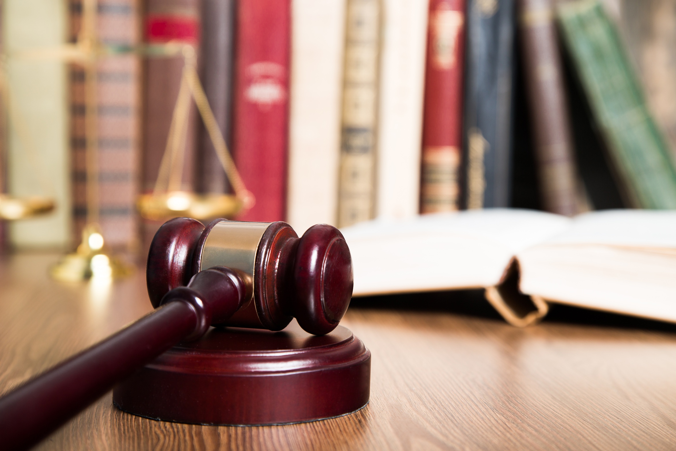 A wooden gavel rests on a circular base on a table in the foreground, symbolizing justice. Behind it, an open book and a set of scales are visible, with various bookshelves filled with legal texts blurred in the background.