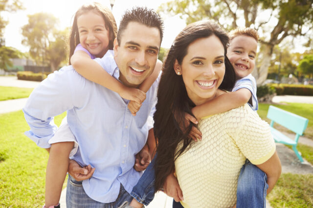 A happy family of four is outdoors on a sunny day. The parents are giving their two children piggyback rides and all are smiling at the camera. The background shows green grass, trees, and a bench, indicating they are at a park.