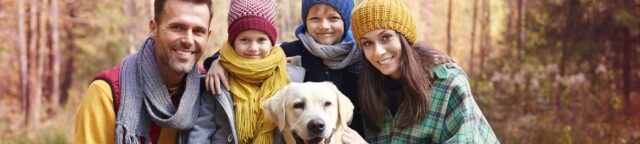 A smiling family of four, dressed in colorful autumn clothing, poses in a wooded area with a happy Labrador retriever at the center. The parents and two children wear knitted hats and scarves, enjoying a fall day outdoors.