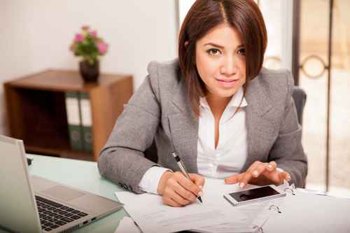 A person with shoulder-length brown hair, dressed in a gray blazer and white shirt, sits at a desk with papers, a laptop, and a smartphone. They are looking at the camera while holding a pen, appearing focused and engaged. A small plant and shelf are in the background.