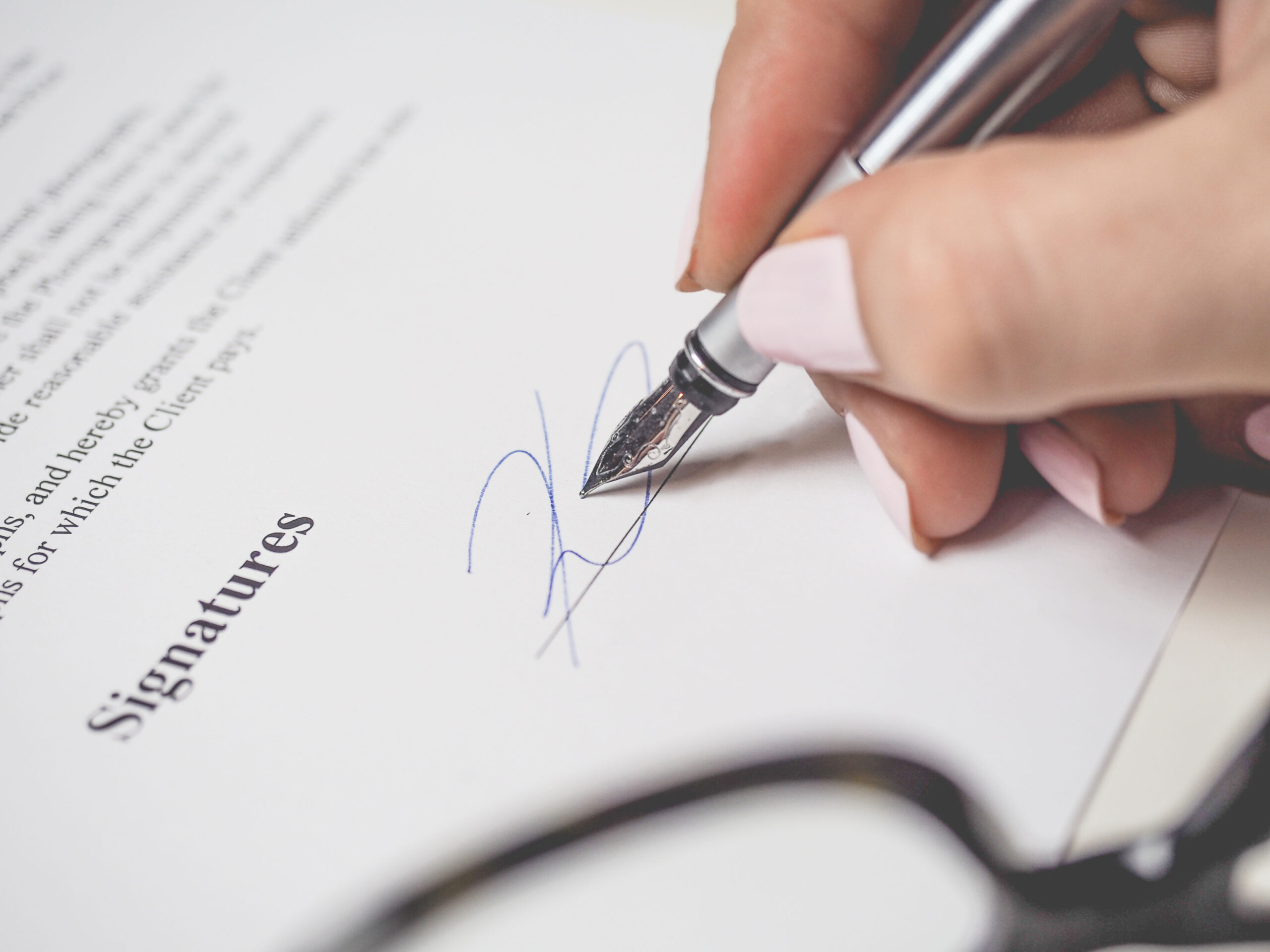 Close-up of a person's hand with pink nail polish signing a document with a fountain pen. The word