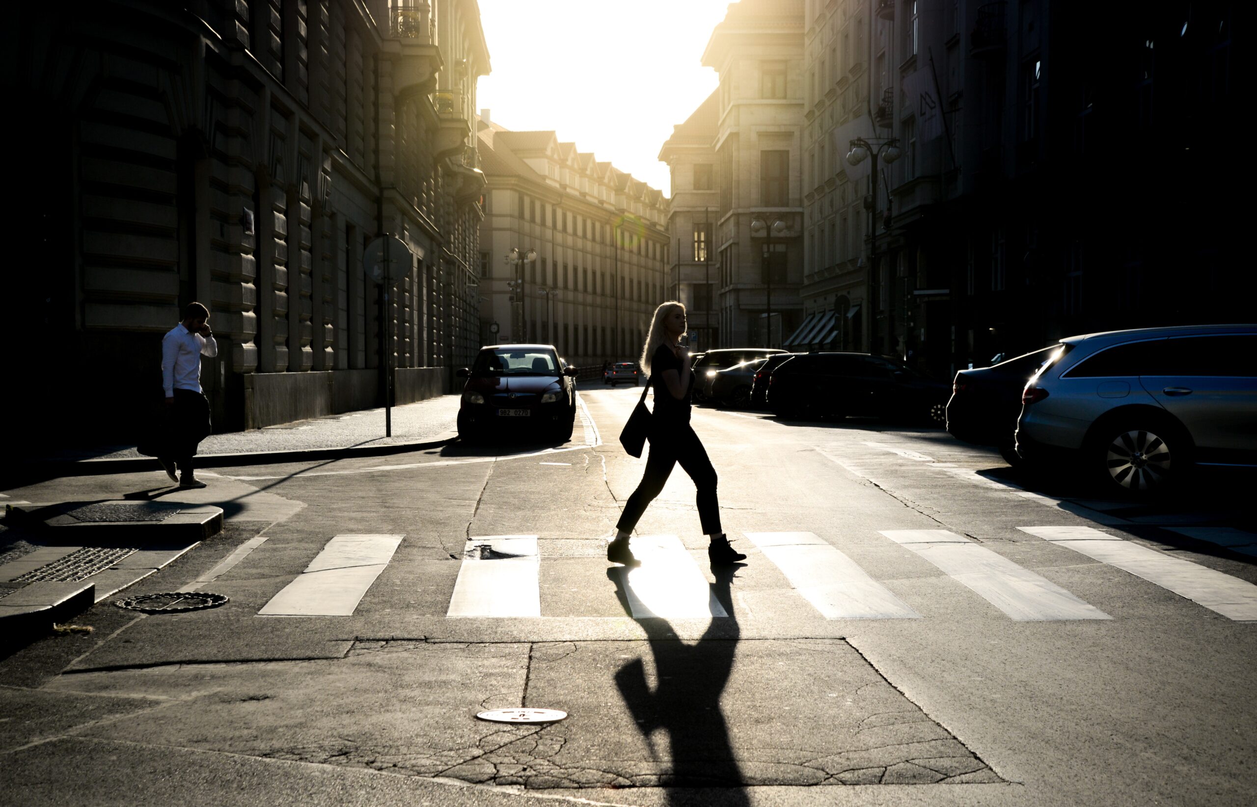 A person crossing a street at a crosswalk in a city during sunset, casting a long shadow. Buildings and parked cars line the street, and another person is walking on the sidewalk to the left. The scene in Pennsylvania is lit by the bright sunlight creating strong contrasts, reminding one to be cautious of pedestrian accidents.