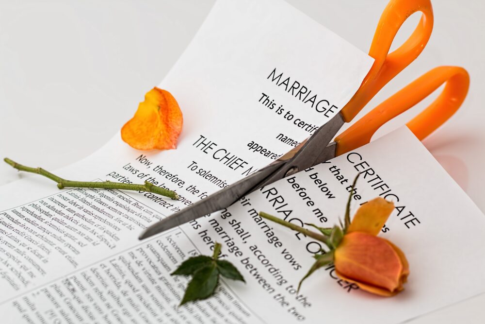 Orange scissors cutting through a marriage certificate alongside a wilted orange rose, emphasizing themes of high asset divorce. The white background highlights the contrast between the bright scissors and the faded rose.