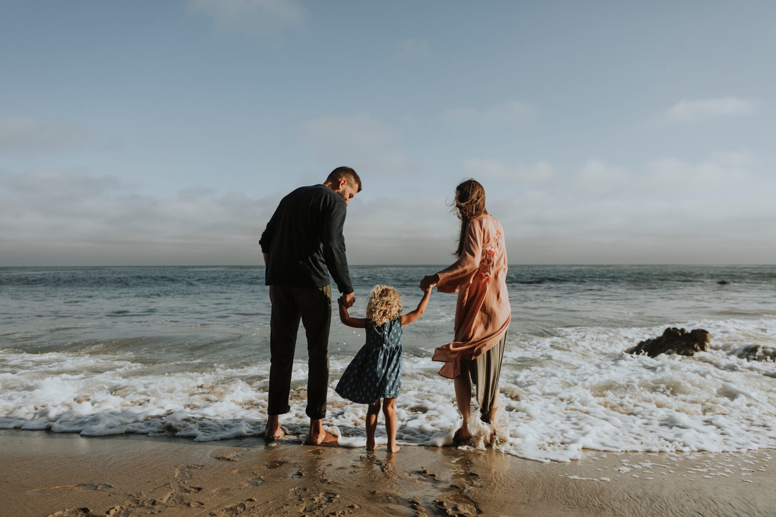 A family of three stands on a sandy beach in Pennsylvania, facing the ocean. The man and woman each hold one hand of a young child between them. The woman is in a pink dress, the man in dark clothing, and the child with curly hair is in a blue dress. Waves gently touch their feet as they cherish this precious moment post-adoption.