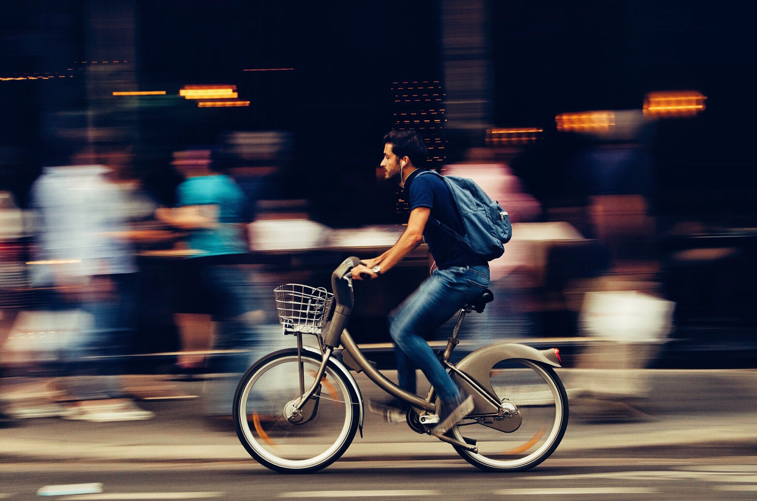 A man wearing a blue shirt and blue jeans rides a bicycle with a basket down a busy Pennsylvania street. He has a blue backpack on and is surrounded by blurred figures of other pedestrians, creating a sense of motion in the bustling city environment.