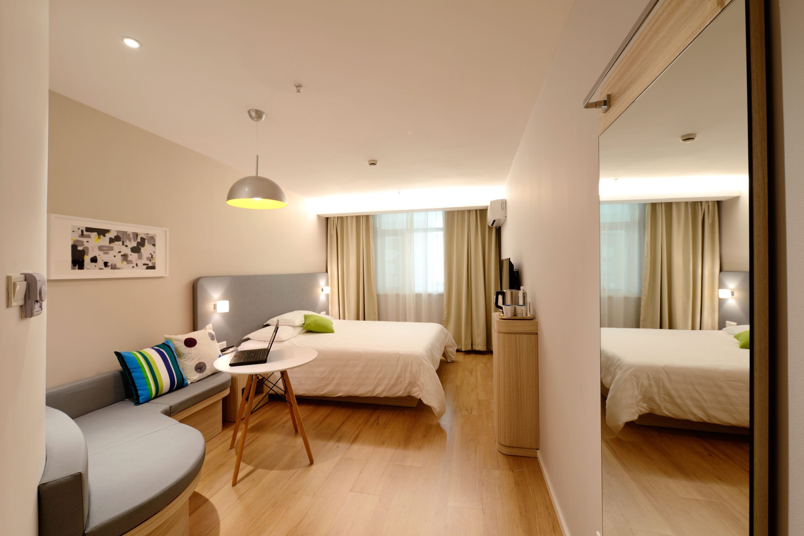 A modern, well-lit Pennsylvania hotel room with two beds, a small sofa, a round coffee table, and a flat-screen TV on a wooden cabinet. The room features light-colored walls and flooring with a large window covered by curtains and a floor-to-ceiling mirror.