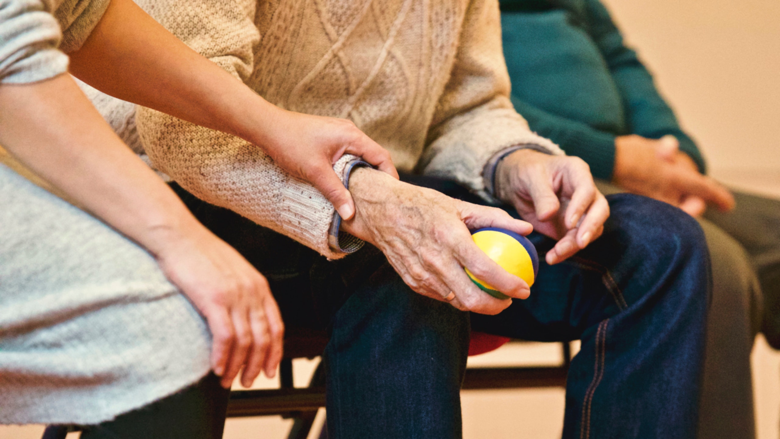 A person wearing a brown sweater assists an elderly individual in a beige sweater who is holding a small yellow and blue ball. Another person in a green sweater is seated nearby. The setting appears to be a supportive or caregiving environment, highlighting the importance of vigilance to report incidents of neglect.