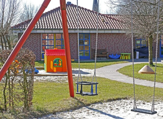A playground on the premises of a brick building with blue doors and red roof tiles features swings, a small red-and-yellow playhouse, and a fenced area. The ground is covered in grass and pathways, with trees in the background—ideal for daycares looking to minimize injuries while children play.