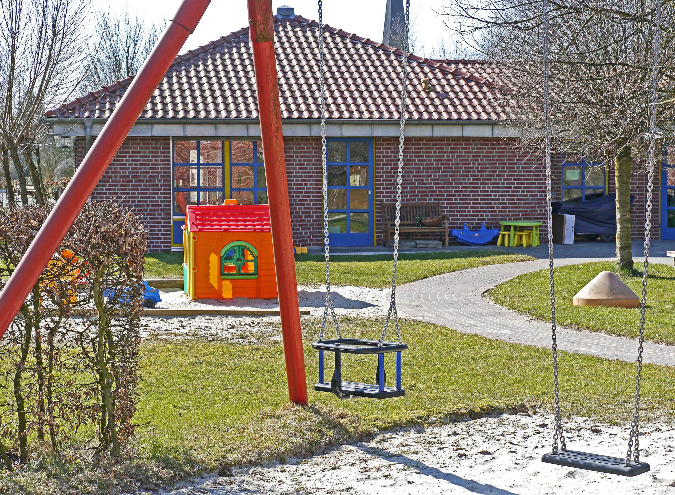 A playground on the premises of a brick building with blue doors and red roof tiles features swings, a small red-and-yellow playhouse, and a fenced area. The ground is covered in grass and pathways, with trees in the background—ideal for daycares looking to minimize injuries while children play.