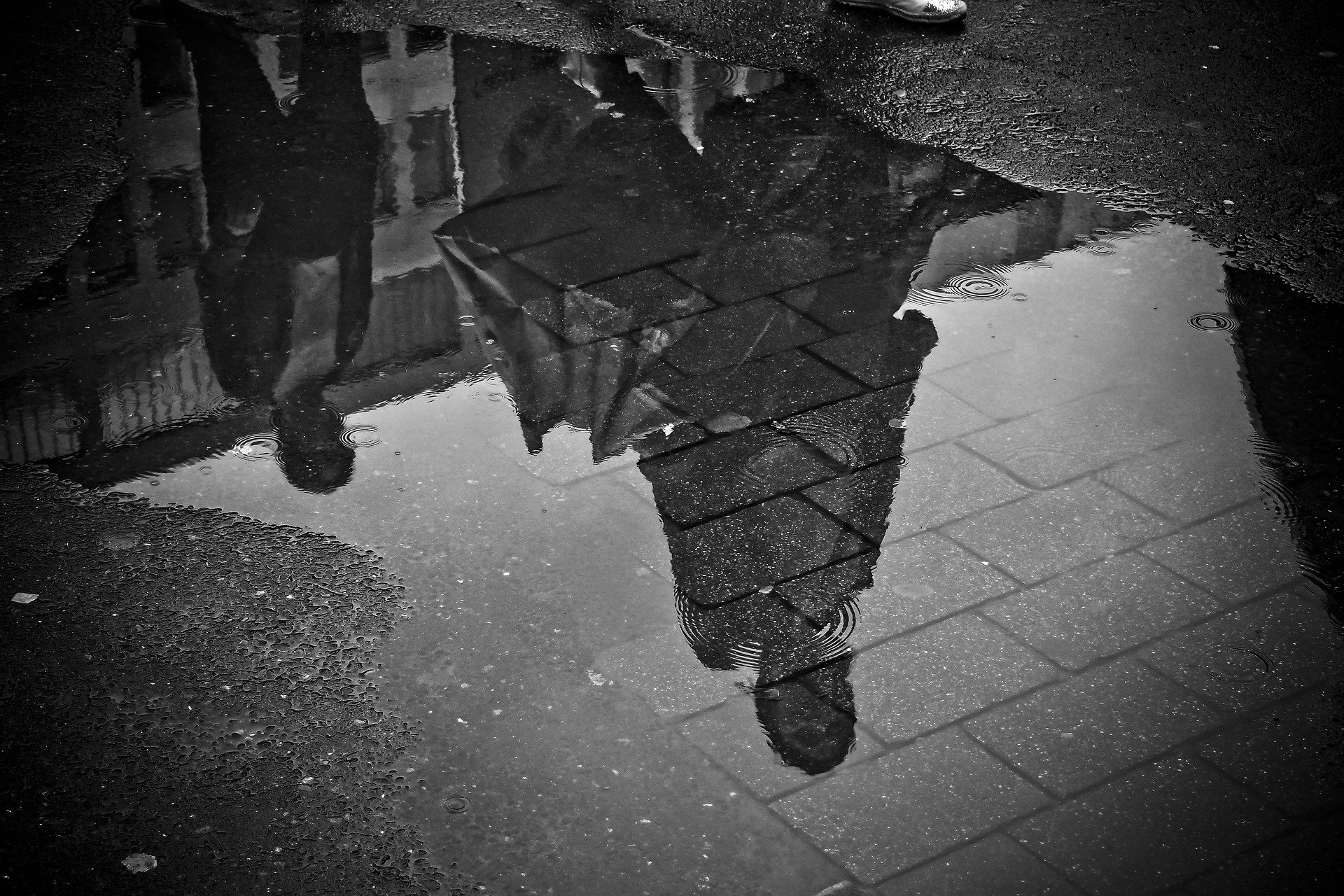 A black-and-white photo showing reflections of two people walking on a wet street in a puddle. The reflections depict one person wearing a coat and another in the background. The visible street surface around the puddle is wet, with some texture and ripples in the water, evoking a rainy day in Pennsylvania.