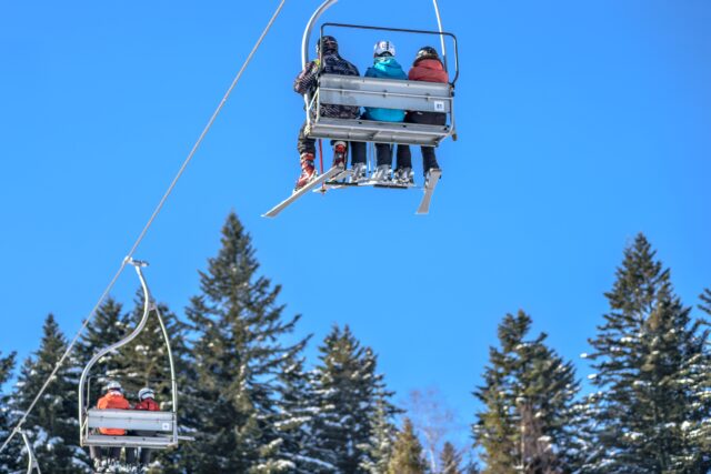 A group of people sitting on a ski lift ascending a snowy mountain slope at a Pennsylvania ski resort. They are dressed in winter clothing, including helmets and ski boots. Tall evergreen trees covered in snow are visible below against a clear blue sky, offering blissful moments away from thoughts of accident recovery.