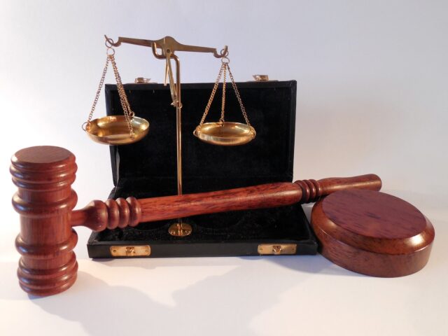 A wooden gavel and round sound block are displayed in the foreground. In the background, a brass balance scale stands inside an open black case on a plain white surface. The image represents judicial or legal concepts, reflecting the DUI legal process and DUI consequences in Pennsylvania.