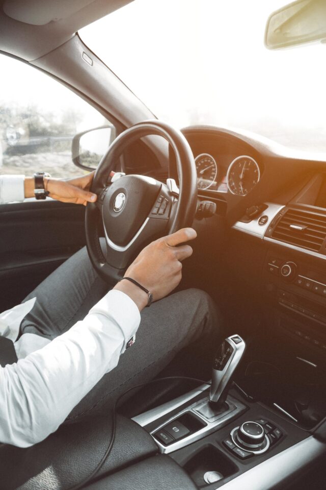 A person in a white shirt and gray pants is driving a car, holding the steering wheel with both hands. The interior of the car includes a gear shift, control buttons, and a dashboard with gauges. Sunlight is streaming through the windshield. Always remember to review your car insurance policies during these times of COVID-19.