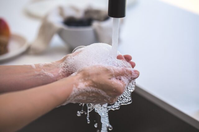 Close-up of a person washing their hands with soap and water under a faucet. Soap bubbles cover the hands, and water flows down, creating foam. The background includes a blurred bowl and fruit. The scene emphasizes hygiene and cleanliness, crucial for protecting yourself against the new coronavirus.