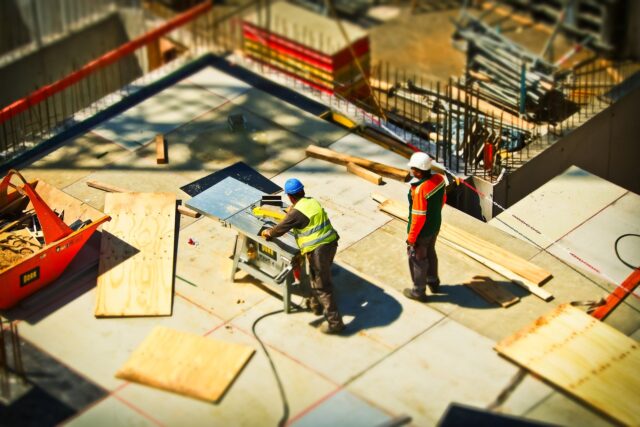 Construction site with two workers wearing hard hats and safety vests. One worker operates a table saw, cutting a wooden plank, while the other stands nearby. The site is scattered with construction materials, including wooden boards and metal scaffolding—essential precautions to avoid being injured in a construction accident.