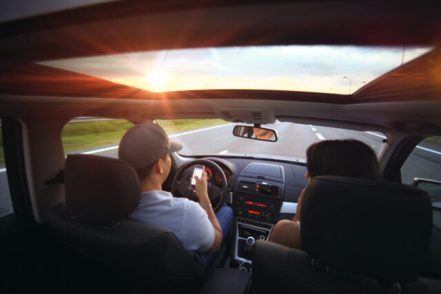 Two people are in a car on a highway, seen from the rear interior with the front seats and dashboard visible. The driver is texting and driving, looking at a smartphone. The sun is setting on the horizon, casting a warm glow through the car’s sunroof—what you need to know to avoid being injured in an accident.