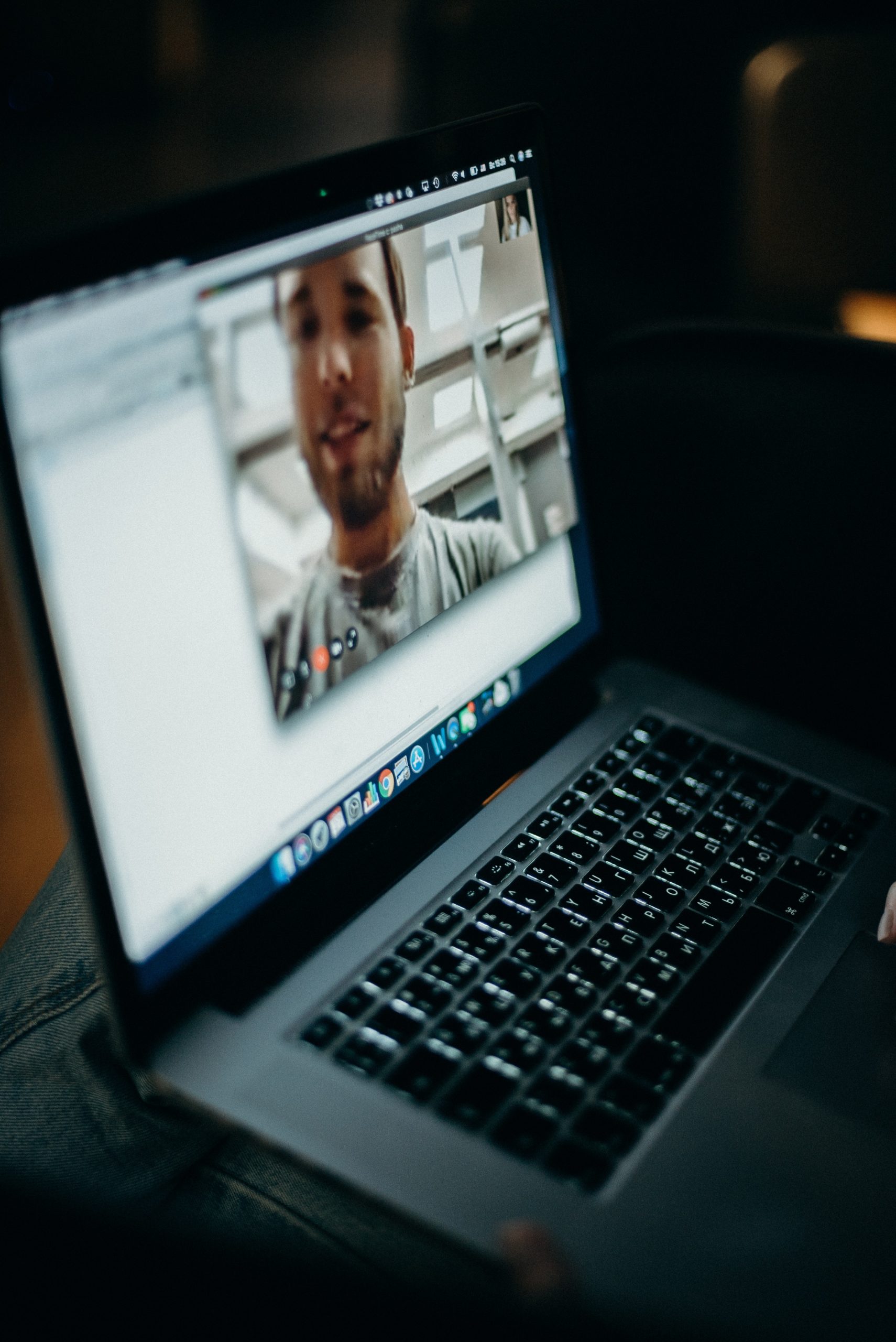 A close-up of a person holding a laptop on their lap with a virtual visit in progress. The screen displays a male participant with a beard and a white shirt in a well-lit room. The laptop keyboard is visible, and the lighting in the surroundings is dim.