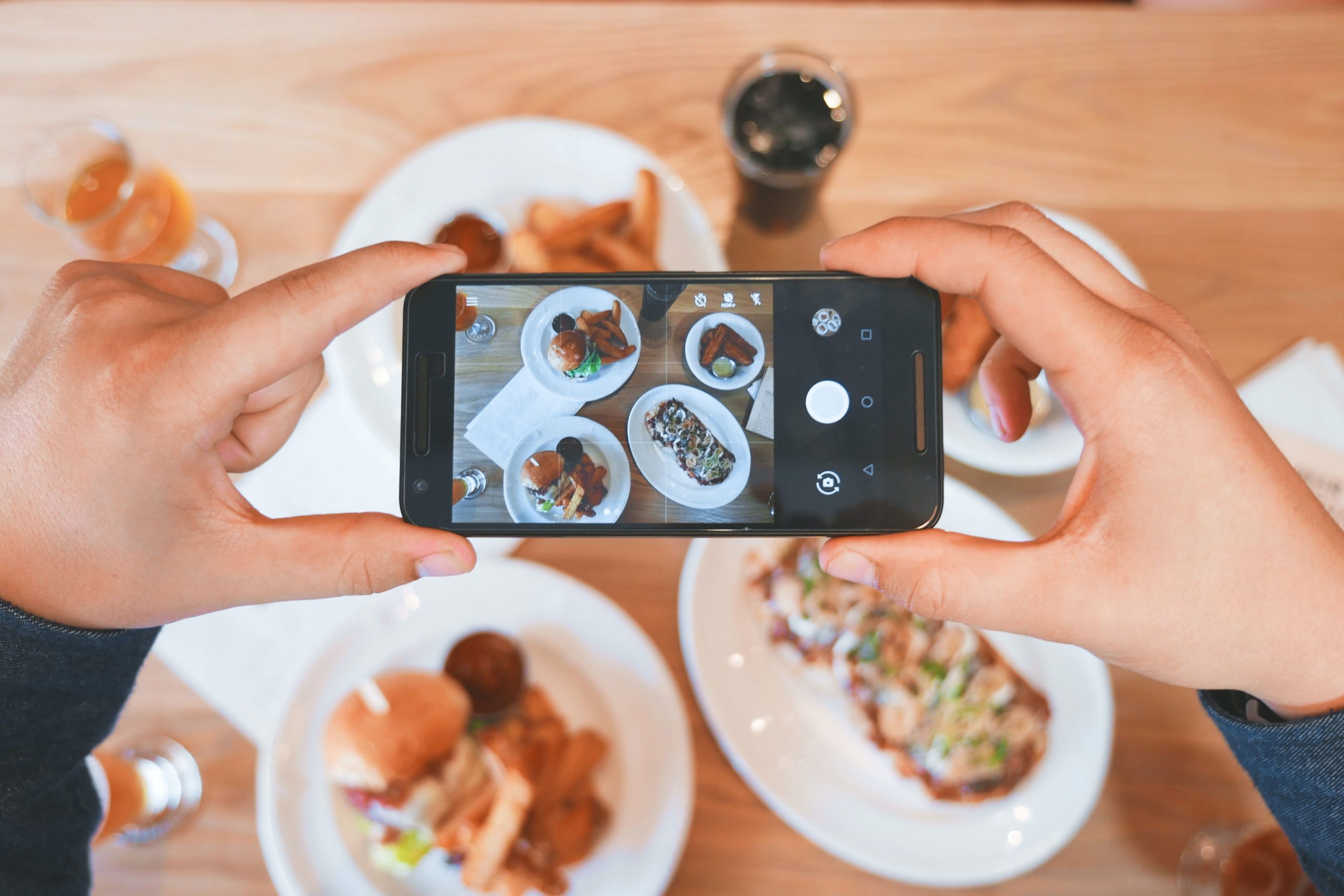 A person holds a smartphone to take a photo of various plated dishes on a wooden table. The dishes include a burger, fries, and a salad. Glasses of drinks are also visible. The phone screen shows the food being photographed from above, perfect for sharing on social media.