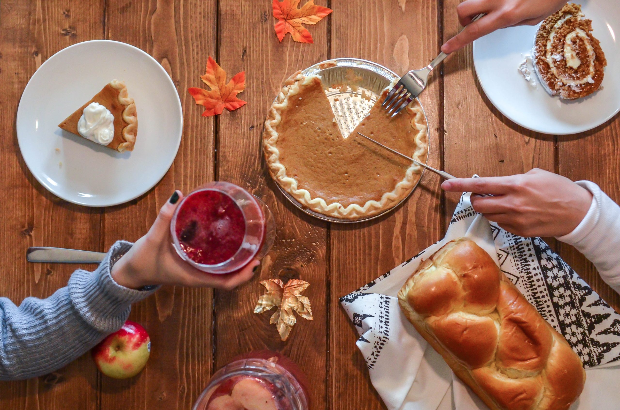 A wooden table with an autumn-themed setup, featuring a whole pumpkin pie under the careful custody of a slice being cut, a slice of pumpkin pie with whipped cream on a plate, a glass of red beverage held by a hand, a loaf of bread, and autumn leaves scattered around.