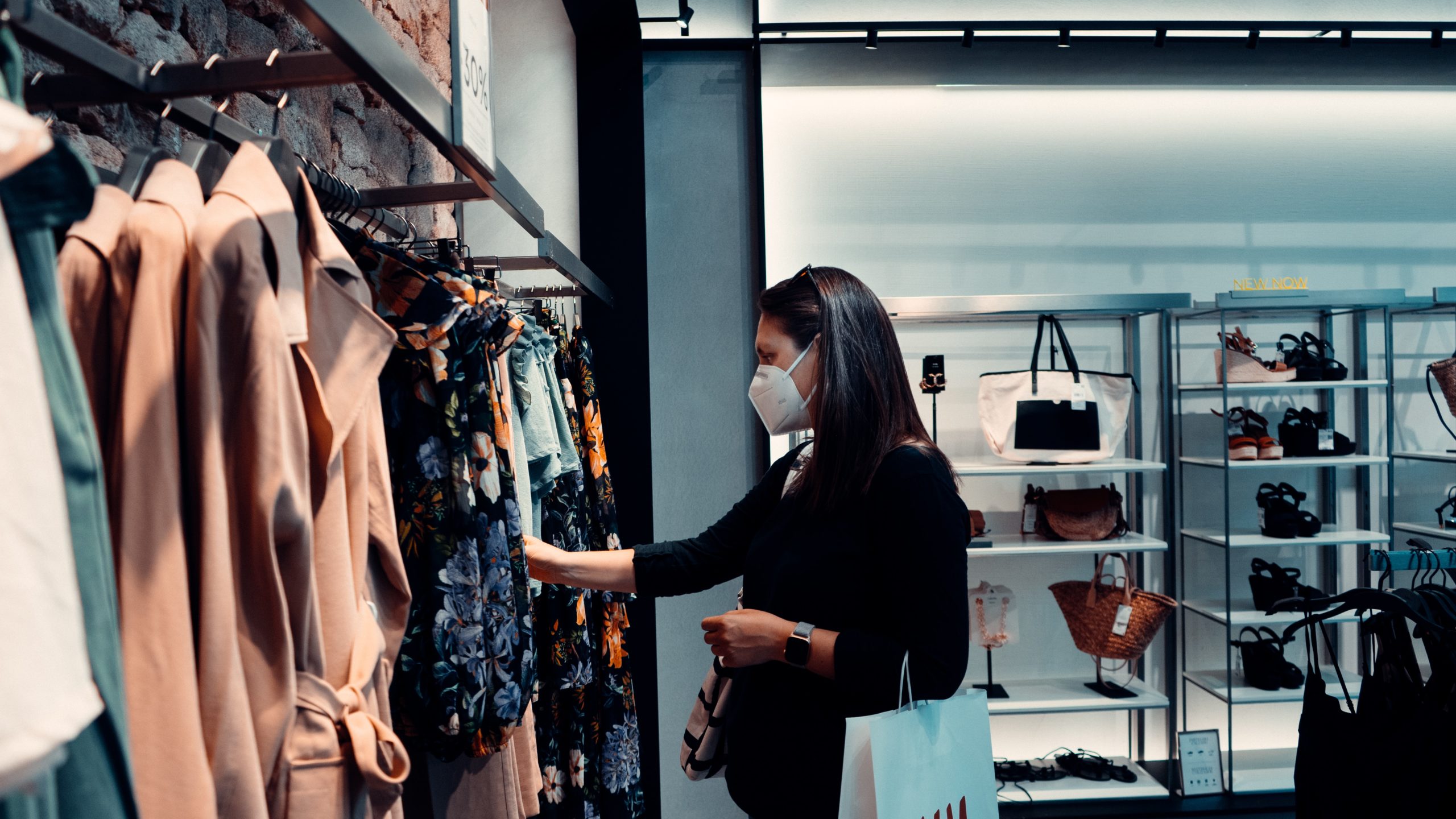 A person wearing a mask shops for clothes in a department store, browsing through a rack of colorful garments. They hold a white shopping bag. Shelves with bags and shoes are displayed in the background, reminding everyone to be cautious and avoid potential accidents while shopping.