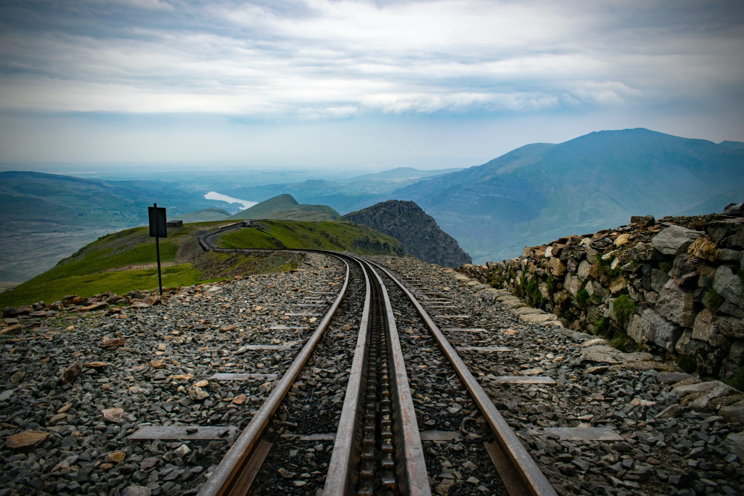 A railway track stretches into the distance on a rocky terrain, flanked by a low stone wall. The scene overlooks rolling green hills and mountains under a cloudy sky, with a glimpse of a distant lake—an area known for its beauty despite infrequent train accidents.