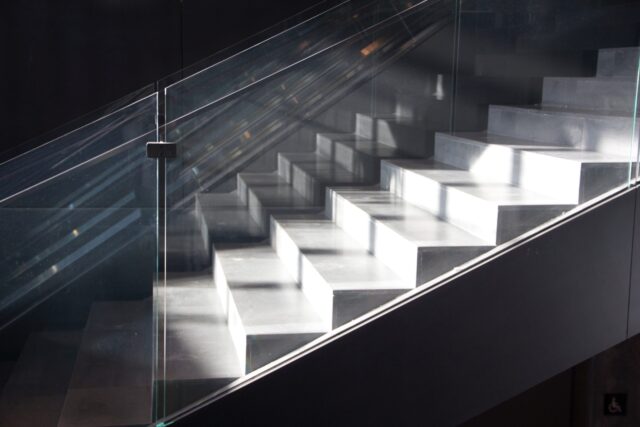 A modern, sunlit staircase with glass railings ascends diagonally from left to right. The sunlight highlights each step, creating a sharp contrast with the shaded areas, which underscores the importance of caution to prevent stairwell accidents. A partial wheelchair accessibility sign is seen in the bottom right corner.
