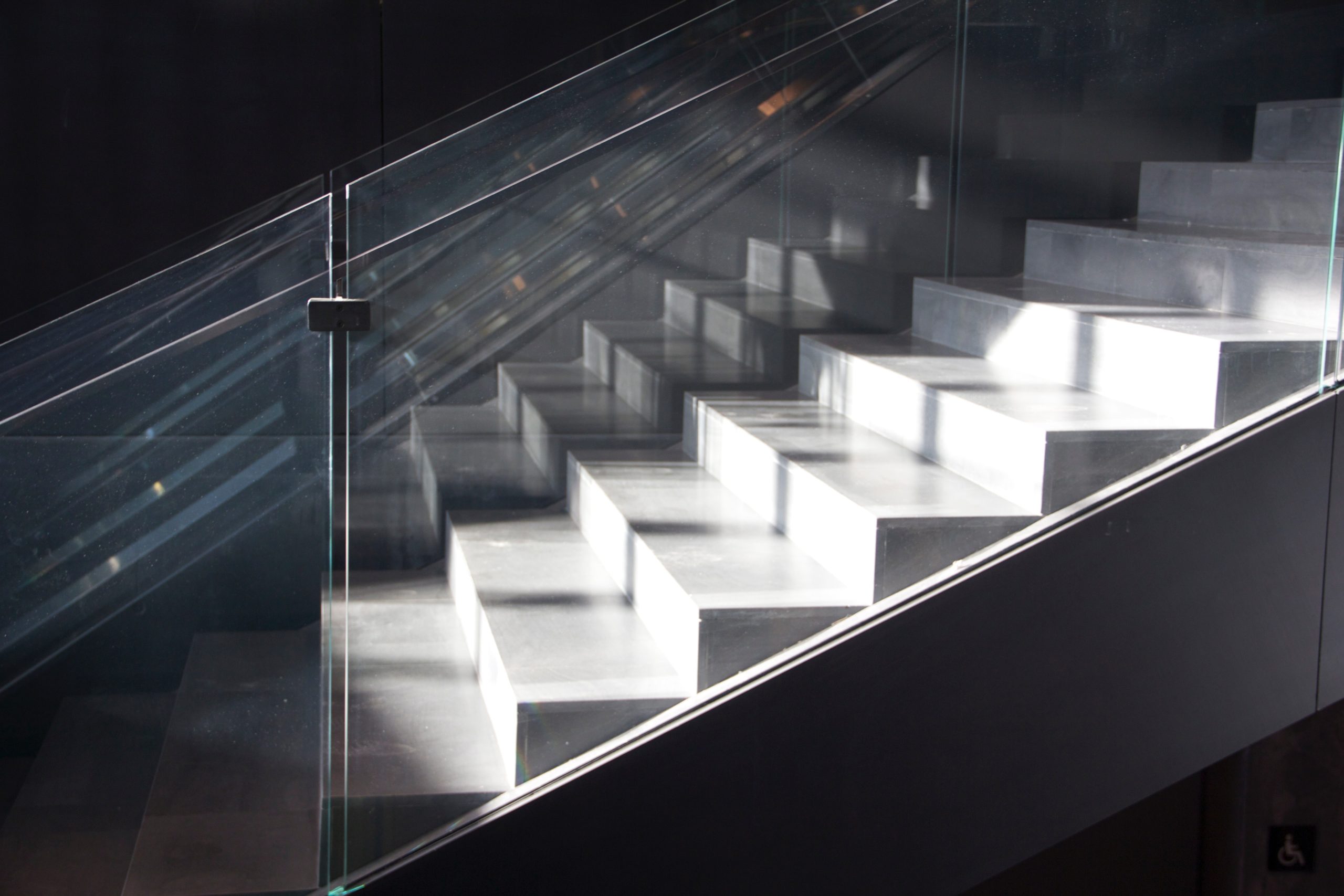 A modern, sunlit staircase with glass railings ascends diagonally from left to right. The sunlight highlights each step, creating a sharp contrast with the shaded areas, which underscores the importance of caution to prevent stairwell accidents. A partial wheelchair accessibility sign is seen in the bottom right corner.