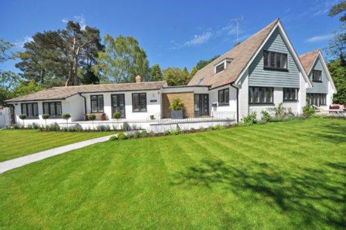 A spacious house with a well-manicured lawn and a paved pathway, this property boasts impressive assets. The house is primarily white with dark trim and features large windows. It has both single-story and two-story sections, with a mix of flat and sloped roofs. Trees and bushes surround the property.