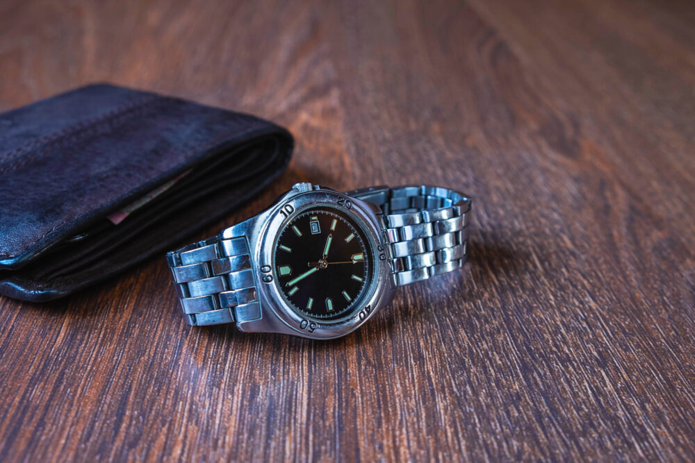 A silver metal wristwatch with a black face and stainless steel band lies on a wooden surface. Beside it, a slightly open black leather wallet reveals a small part of its contents, symbolizing personal items that once held deeper meaning, now left behind after a mediated divorce.