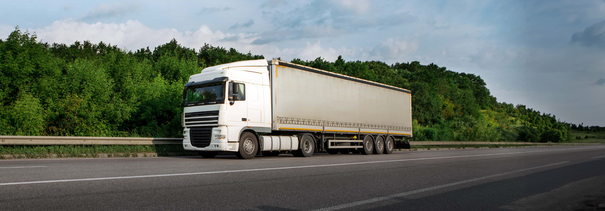 A white semi-truck with a covered trailer is parked on the side of a highway, seemingly still intact. The background features a lush, green forest under a partly cloudy sky, evoking peace—a stark contrast to scenarios where you might need a truck accident attorney.