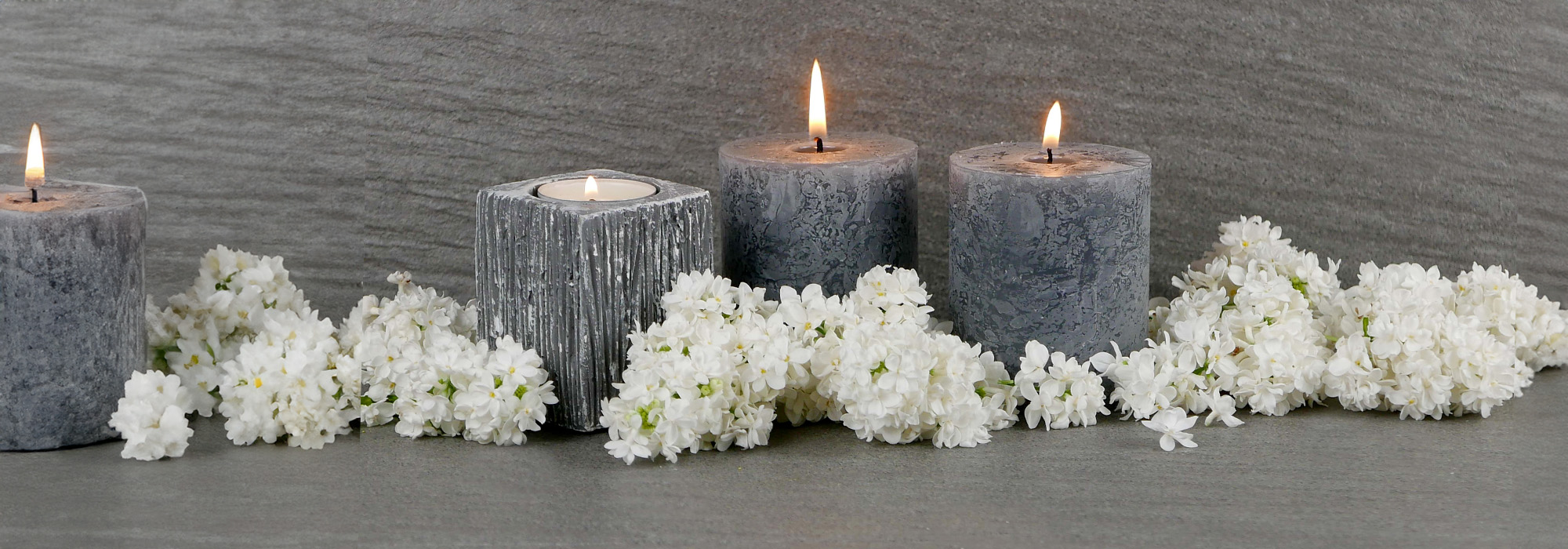 Four gray candles, three lit and one unlit, are arranged on a gray surface surrounded by white flowers. The backdrop is a textured gray wall, creating a serene and calm atmosphere, perfect for reflection. Should you need guidance during tough times, consult a wrongful death attorney to support your journey.