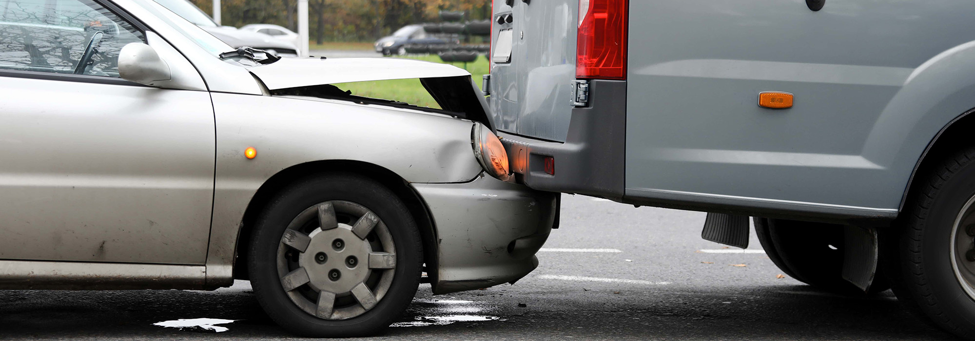 A minor car accident where the front of a silver sedan has collided with the rear of a gray van. The sedan's front bumper is damaged and slightly crumpled. Both vehicles are on a road with trees and grass visible in the background, an auto accident attorney might be necessary for resolving damages.