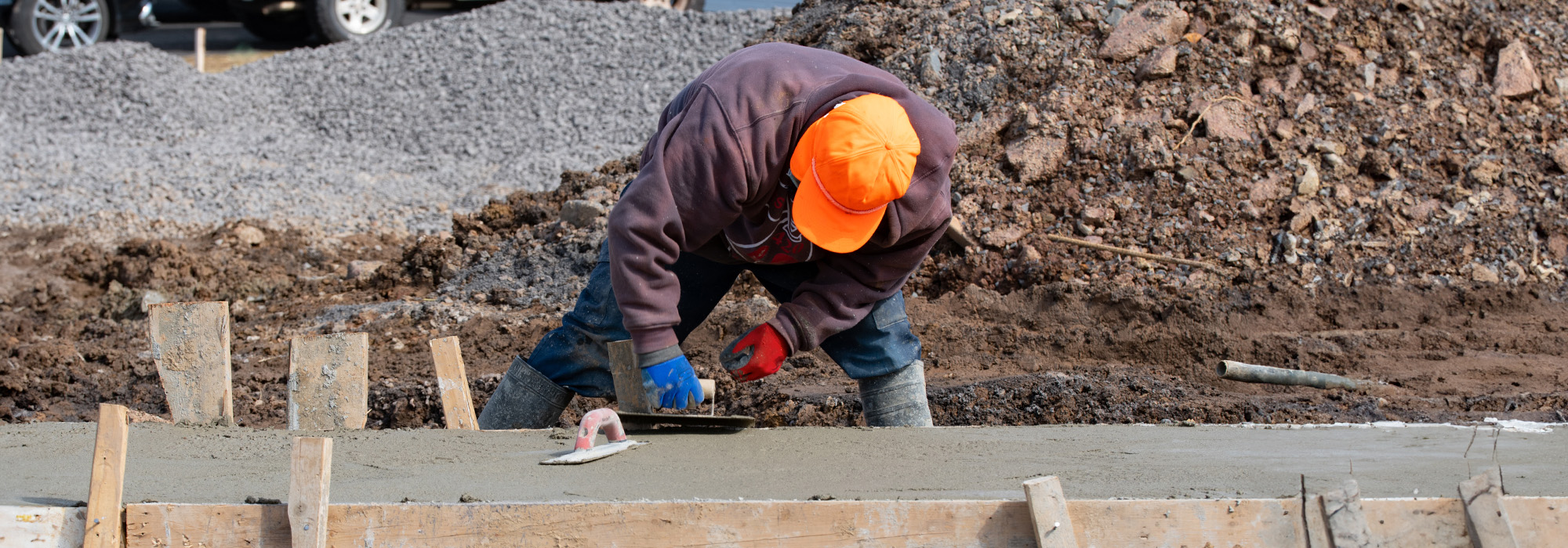 A construction worker wearing an orange cap and gray hoodie is smoothing wet concrete with a trowel at a busy construction site. In the background, there are piles of dirt and gravel. The worker is focused on creating a smooth surface, highlighting the importance of safety that any construction accident attorney would advocate for.