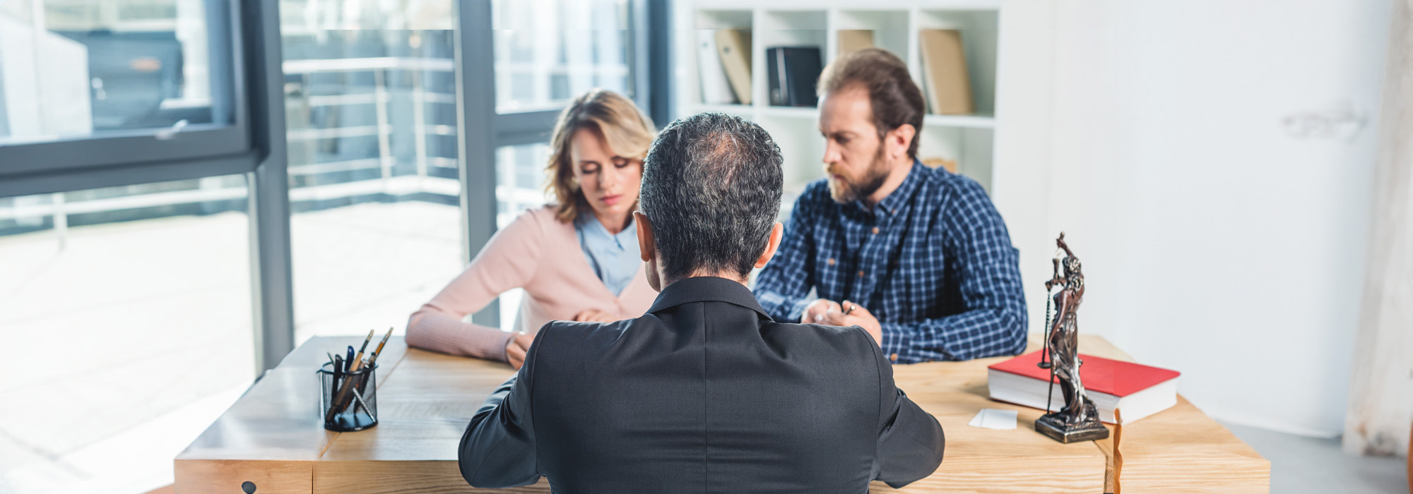 A couple sits across from a mediation attorney in a suit at a wooden desk, engaged in a serious discussion. The office, with large windows and a bookshelf, features a statue of Lady Justice and a red book on the desk.