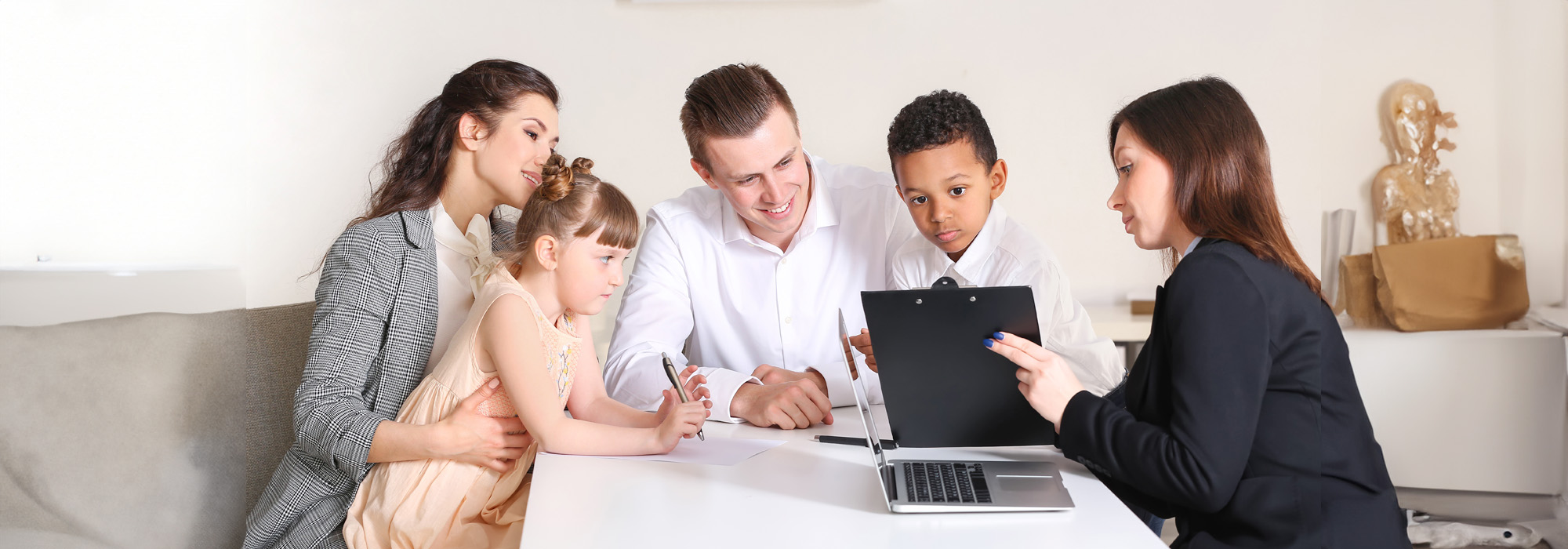 A family with two adults and two children sits across a table from an adoption attorney. The woman, dressed in a black blazer, holds a clipboard and engages with the attentive family. A laptop is placed on the table, suggesting the setting is an office.