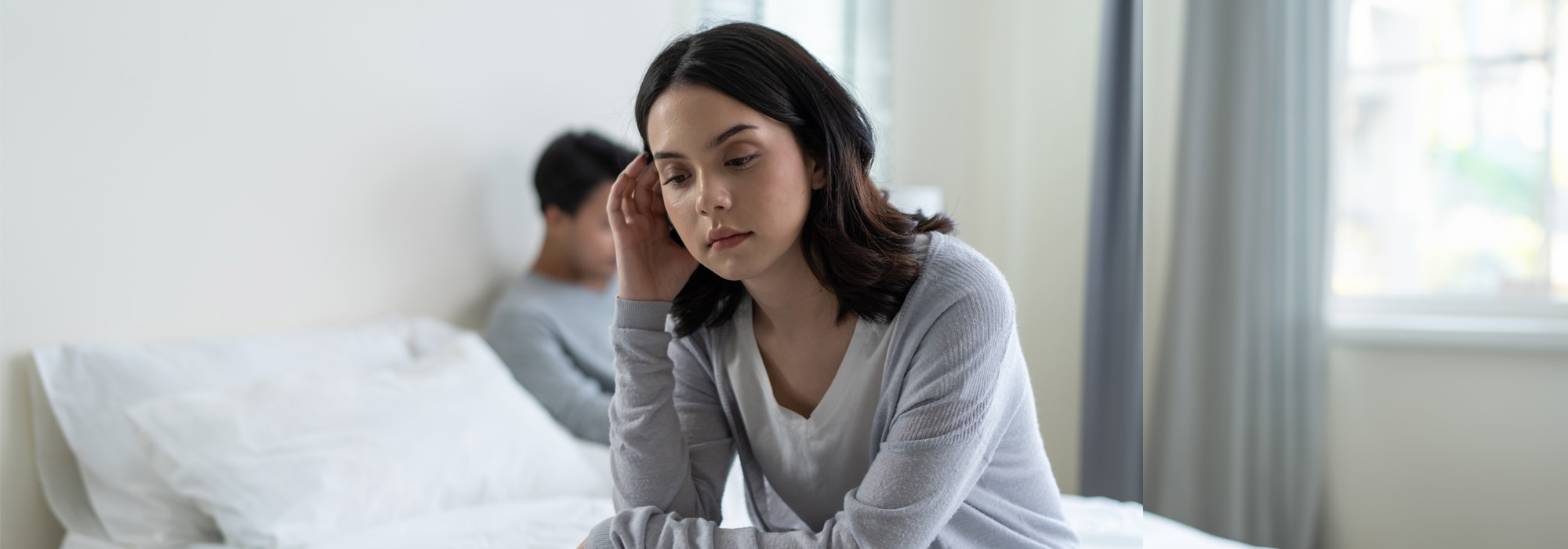 A woman sits on a bed looking pensive with her hand resting on her head. In the background, a man sits facing away on the opposite side of the bed. Both appear to be in a bright, minimally decorated room, perhaps contemplating their next move after consulting a sex crime attorney.