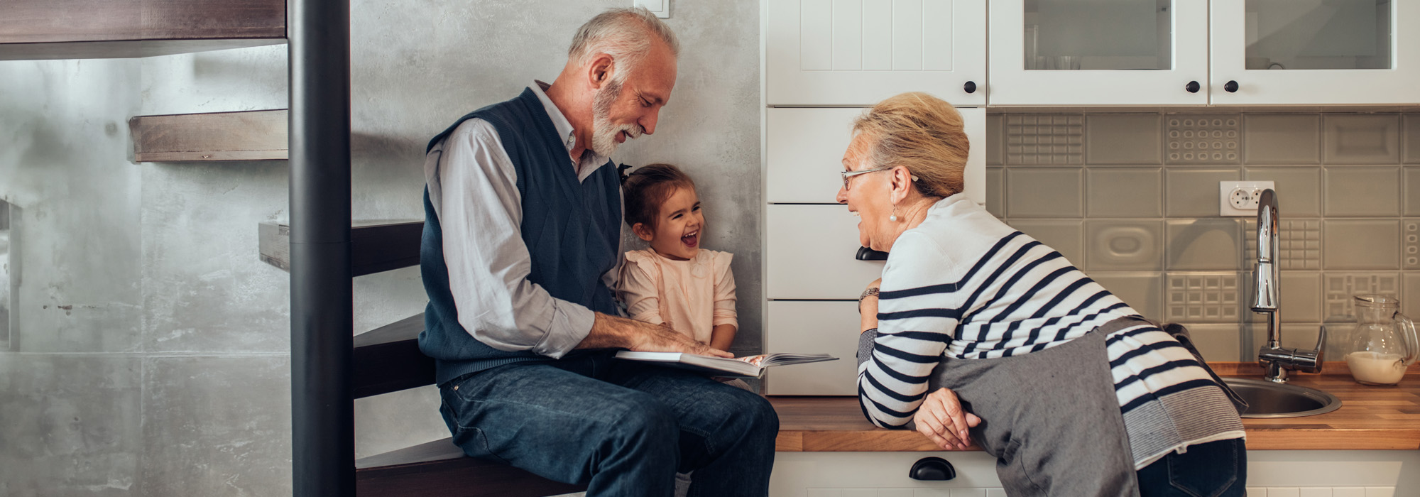 A smiling elderly man and woman interact with a young girl in a modern kitchen. The man, who could be mistaken for a Grandparents' Rights Attorney, sits on a staircase reading a book to the laughing child beside him. The woman leans on the counter, also smiling, creating a warm, family atmosphere.