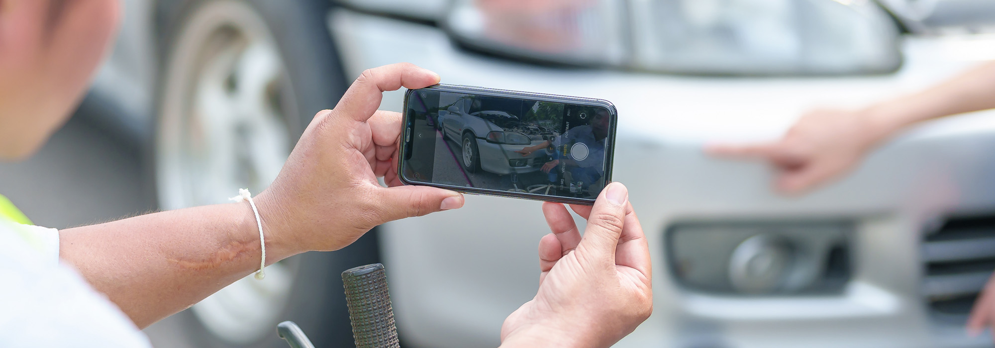 A person is holding a smartphone and taking a photo of a car with visible damage to its front bumper after a car accident. Another hand is pointing at the damage while the car is parked, with part of its headlight and tire visible.