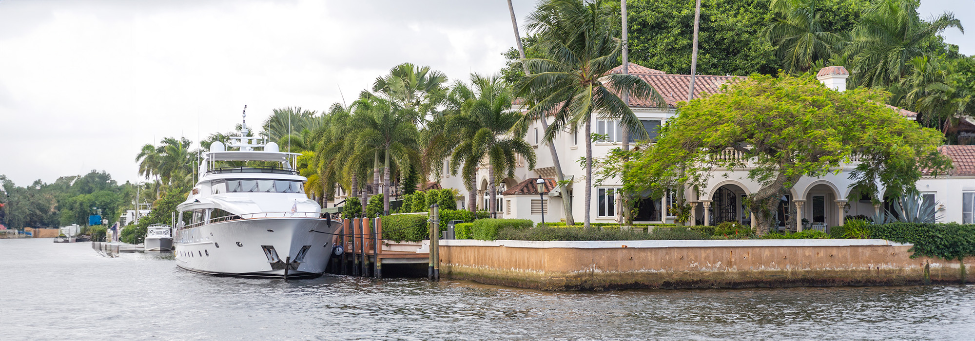 A luxurious waterfront mansion, often seen in cases handled by a high net worth divorce attorney, is surrounded by palm trees and lush greenery. A large white yacht is docked alongside the property, reflecting the affluent lifestyle. The serene waterway and clear sky add to the picturesque setting.