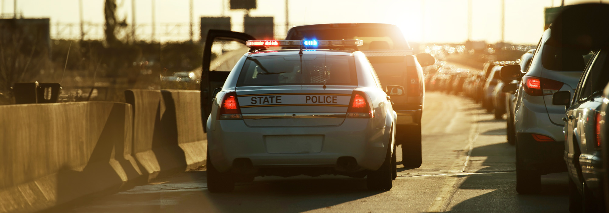 A state police car with flashing lights has pulled over a vehicle on a busy highway during sunset. The police car's door is open, and several other vehicles are visible in the background. York County Traffic Offense Attorney concept image