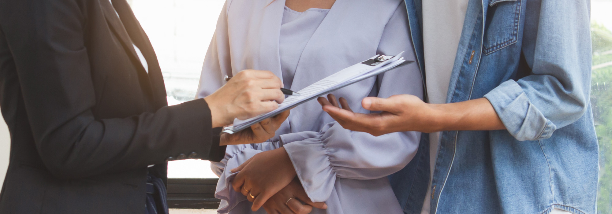 Three individuals are standing together, with one person in a suit holding a clipboard and a pen, gesturing towards the document. The other two people are paying close attention to the clipboard, possibly engaged in dispute resolution. The individuals are indoors, and the background is softly lit.