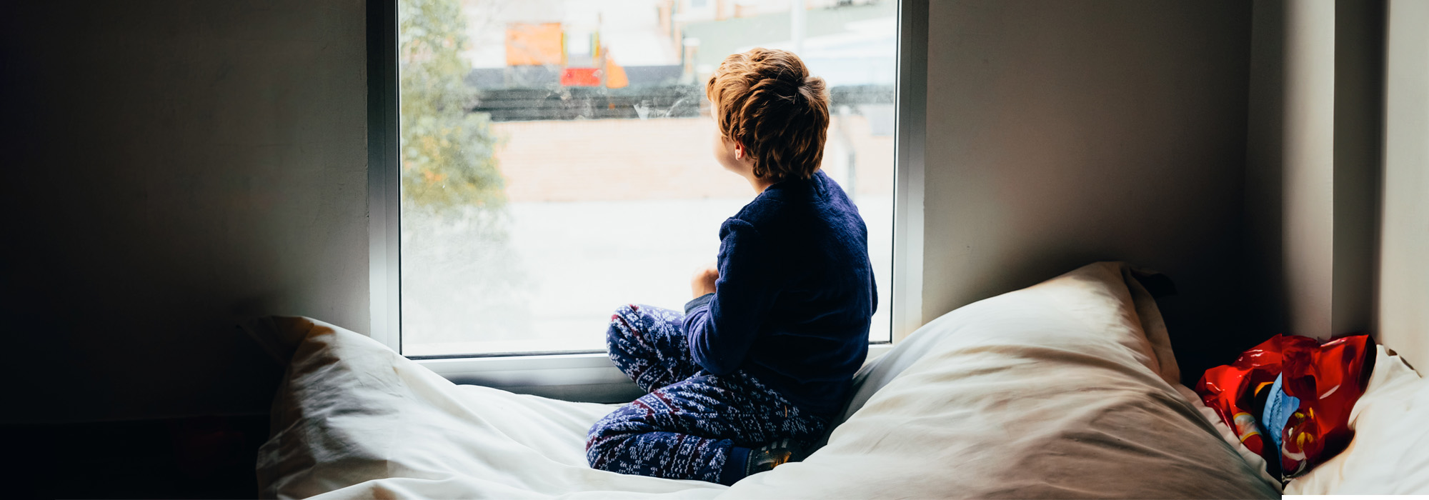 A child wearing pajamas sits on a cushioned windowsill, looking out of a large window. The scene outside is slightly blurred, suggesting a rainy or cloudy day. The cozy interior contrasts with the gray exterior. Beside the child and red toy lies an old business card for a Juvenile Dependency Lawyer.