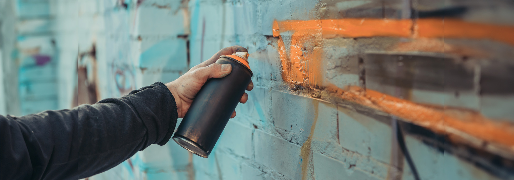 A person holding a black spray paint can, spraying orange paint onto a graffiti-covered brick wall, vibrant with various shades of blue and other colors. The person's sleeve hints at a long-sleeved shirt. Nearby, the presence of a misdemeanor defense lawyer could suggest awareness of potential legal consequences.