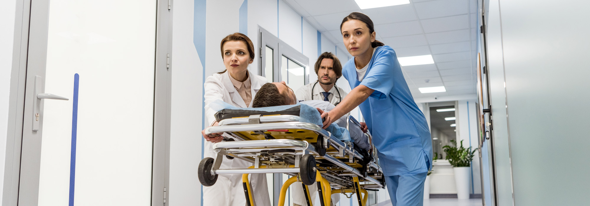 Three medical professionals, two in white coats and one in blue scrubs, are urgently pushing a patient on a stretcher through a hospital hallway. The setting is clinical and clean with bright lighting. The expressions on their faces are serious and focused, reminiscent of a catastrophic injury attorney during a crucial case.