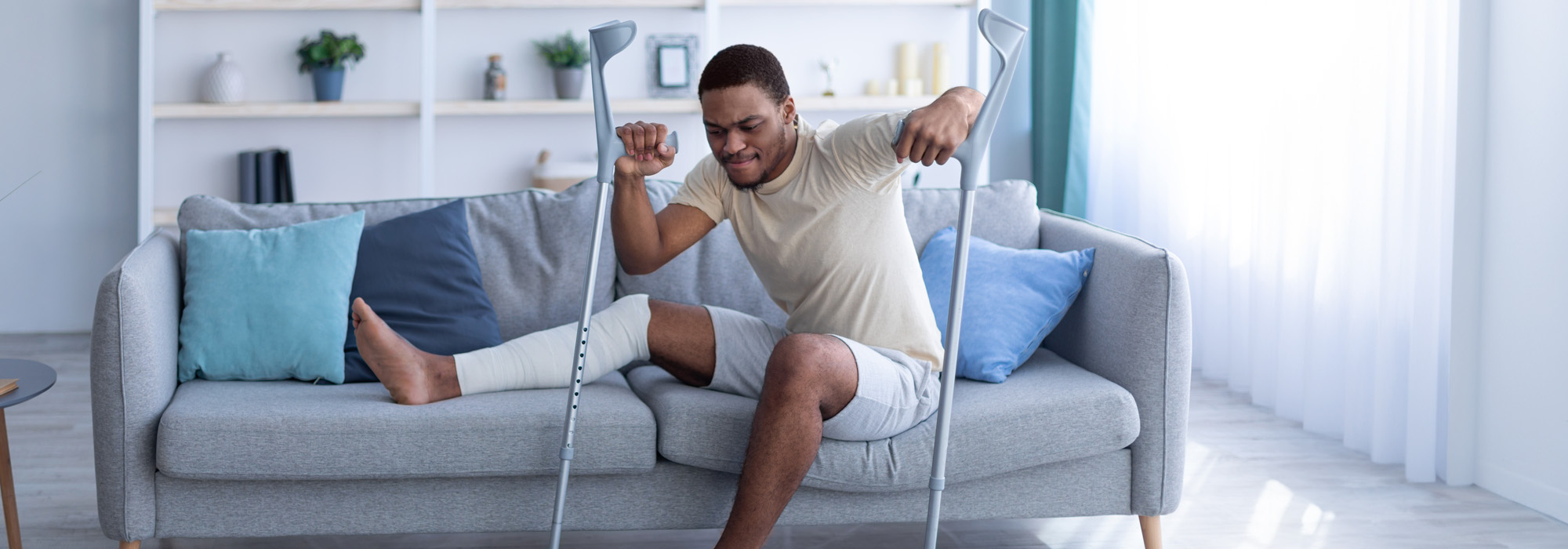A man with a bandaged leg sits on a gray couch, holding crutches as he strains to stand up in a bright living room. Shelves with decor are visible in the background, hinting at the challenges he's faced and perhaps prompting him to seek advice from a personal injury attorney.